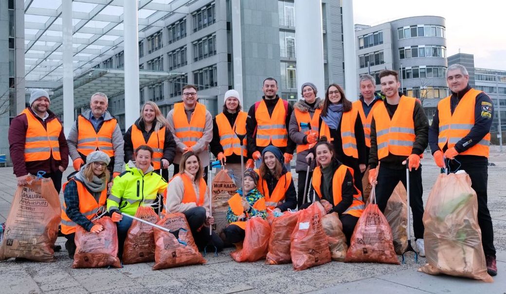 Auf dem Bild sind mehrere Personen mit orangenen Warnwesten, Müllsäcken und Müllgreifer zu sehen. 