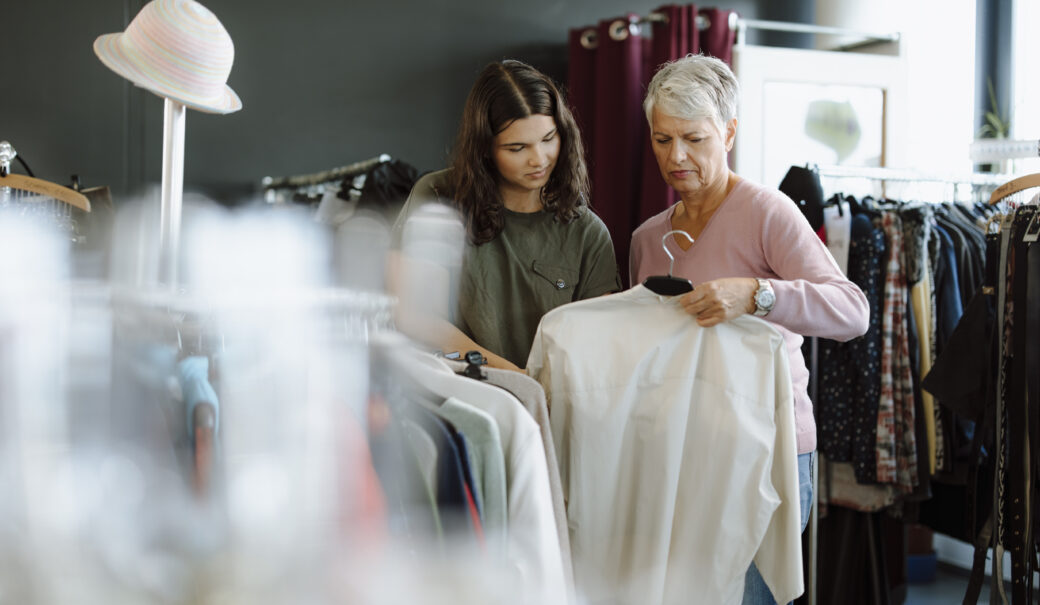 Eine ältere Dame mit grauem Haar steht in einem Secondhandladen und hält eine weiße Bluse in der Hand. Neben ihr steht ein junges Mädchen mit lockigem Haar und einem olivgrünen T-Shirt, das ebenfalls die Bluse interessiert betrachtet.